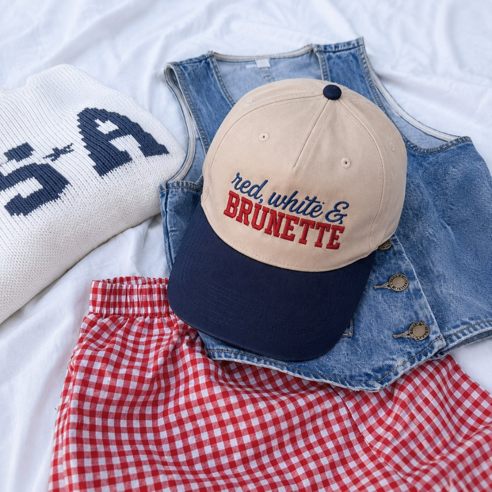 A beige and navy baseball cap with 'red, white & BRUNETTE' embroidered on the front, styled in a flat lay with a white knit sweater, a denim vest, and a red-and-white gingham skirt on white fabric.