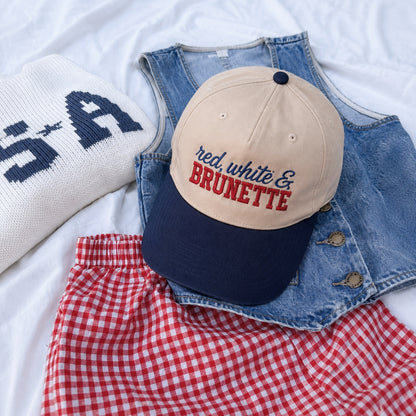 A beige and navy baseball cap with 'red, white & BRUNETTE' embroidered on the front, styled in a flat lay with a white knit sweater, a denim vest, and a red-and-white gingham skirt on white fabric.