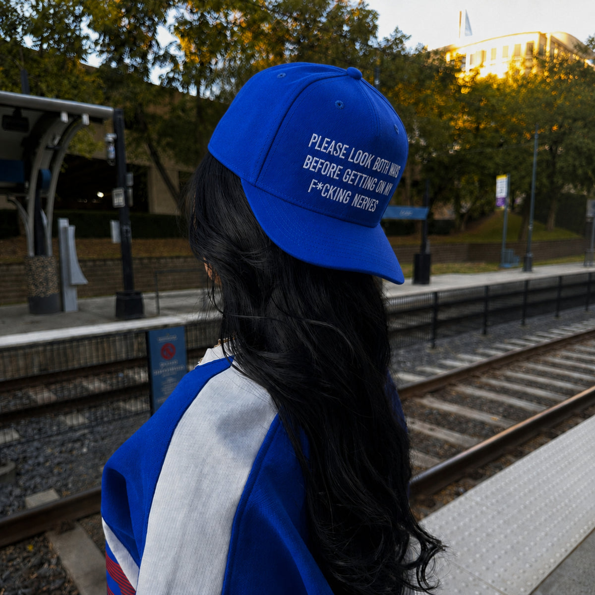 A person standing at a train station platform, shown from the back wearing a bright blue baseball cap with white embroidered text that reads, 'PLEASE LOOK BOTH WAYS BEFORE GETTING ON F*CKING RAILS.'.