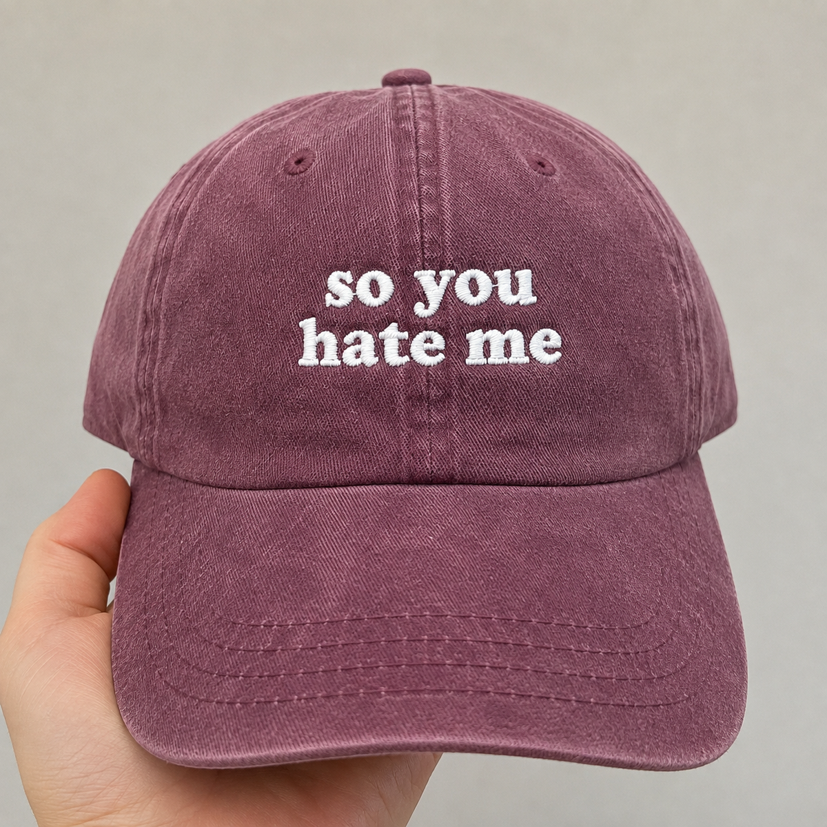 Washed burgundy baseball cap with white embroidered “so you hate me” text, featuring a minimalist sarcastic quote design and vintage distressed style.
