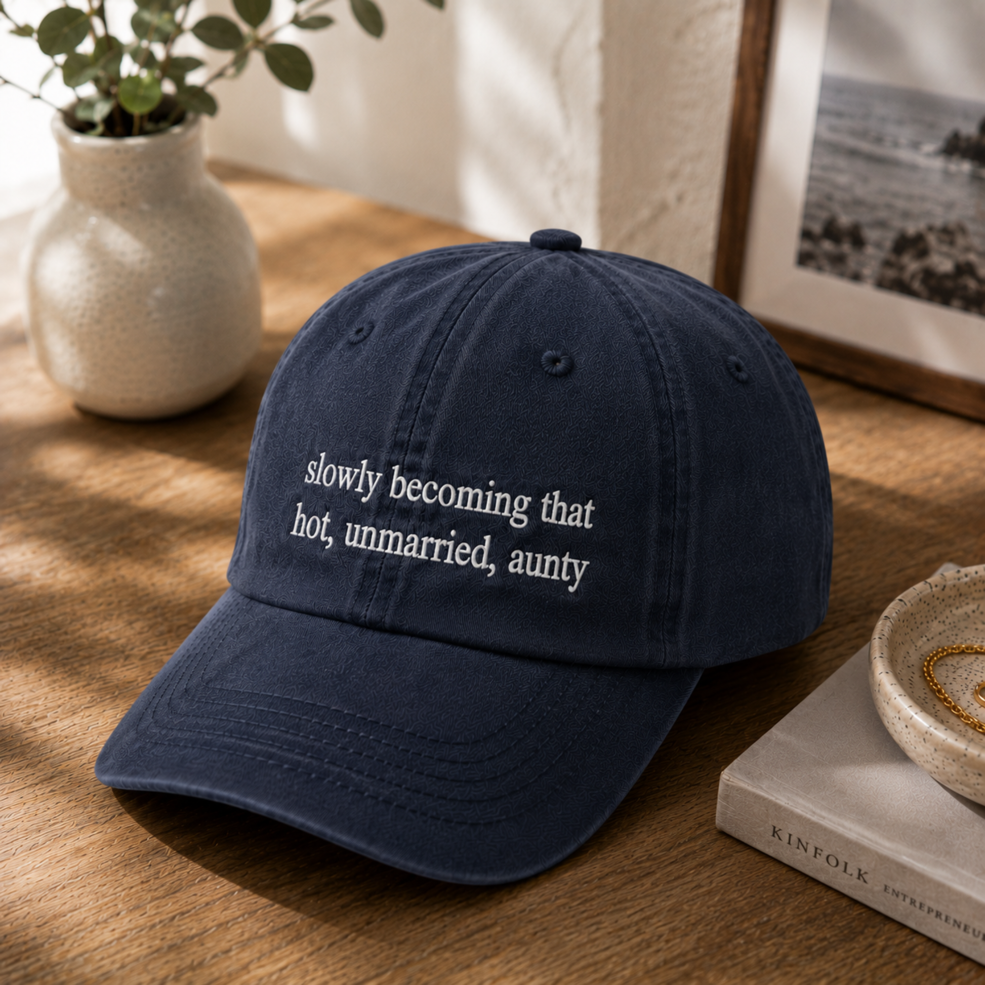 A navy blue dad hat baseball cap with white embroidered text reading 'slowly becoming that hot, unmarried, aunty' resting on a wooden table next to a small vase and a book.