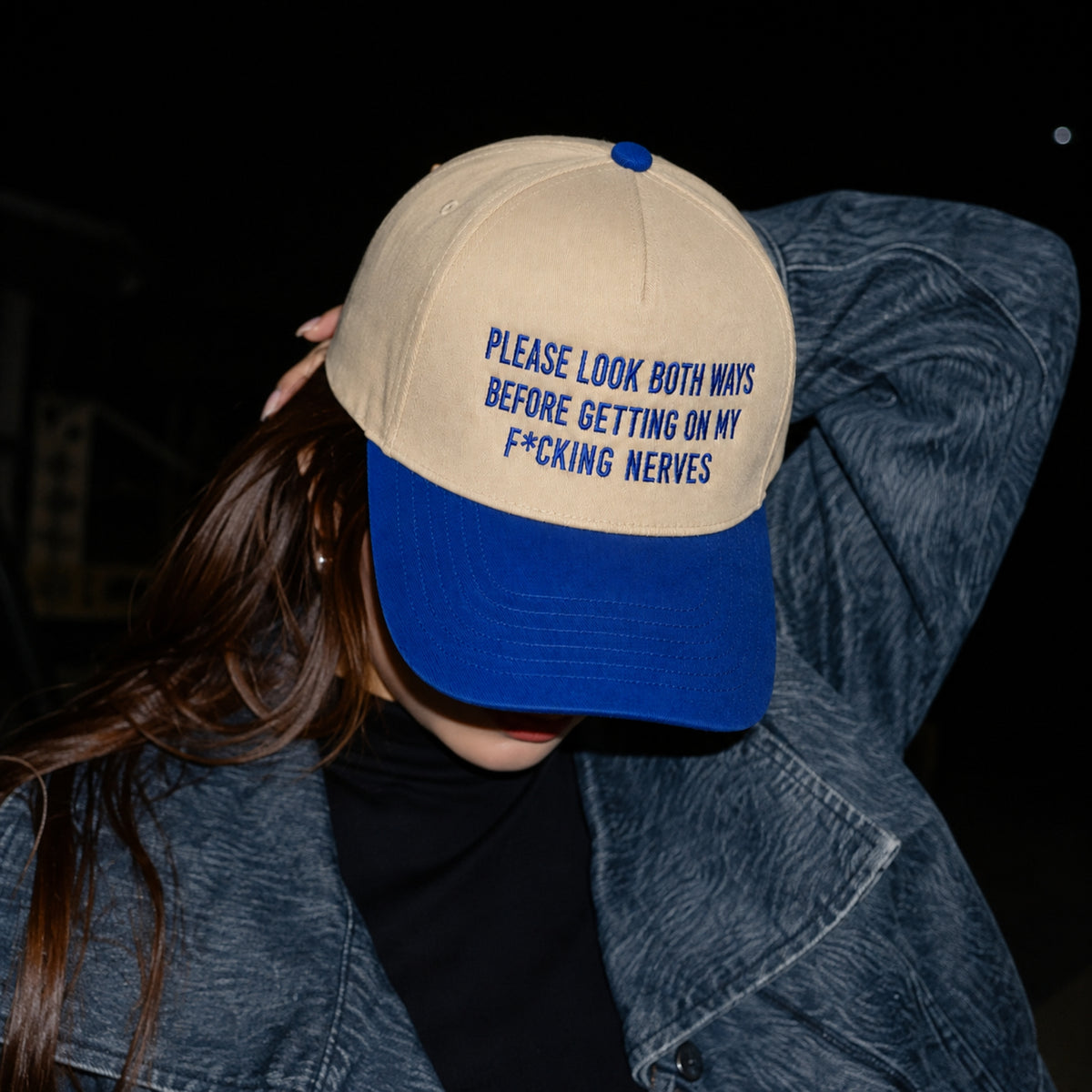 A person wearing a beige and blue baseball cap with blue embroidered text that reads 'PLEASE LOOK BOTH WAYS BEFORE GETTING ON MY F*CKING NERVES', paired with a blue denim jacket against a dark background.