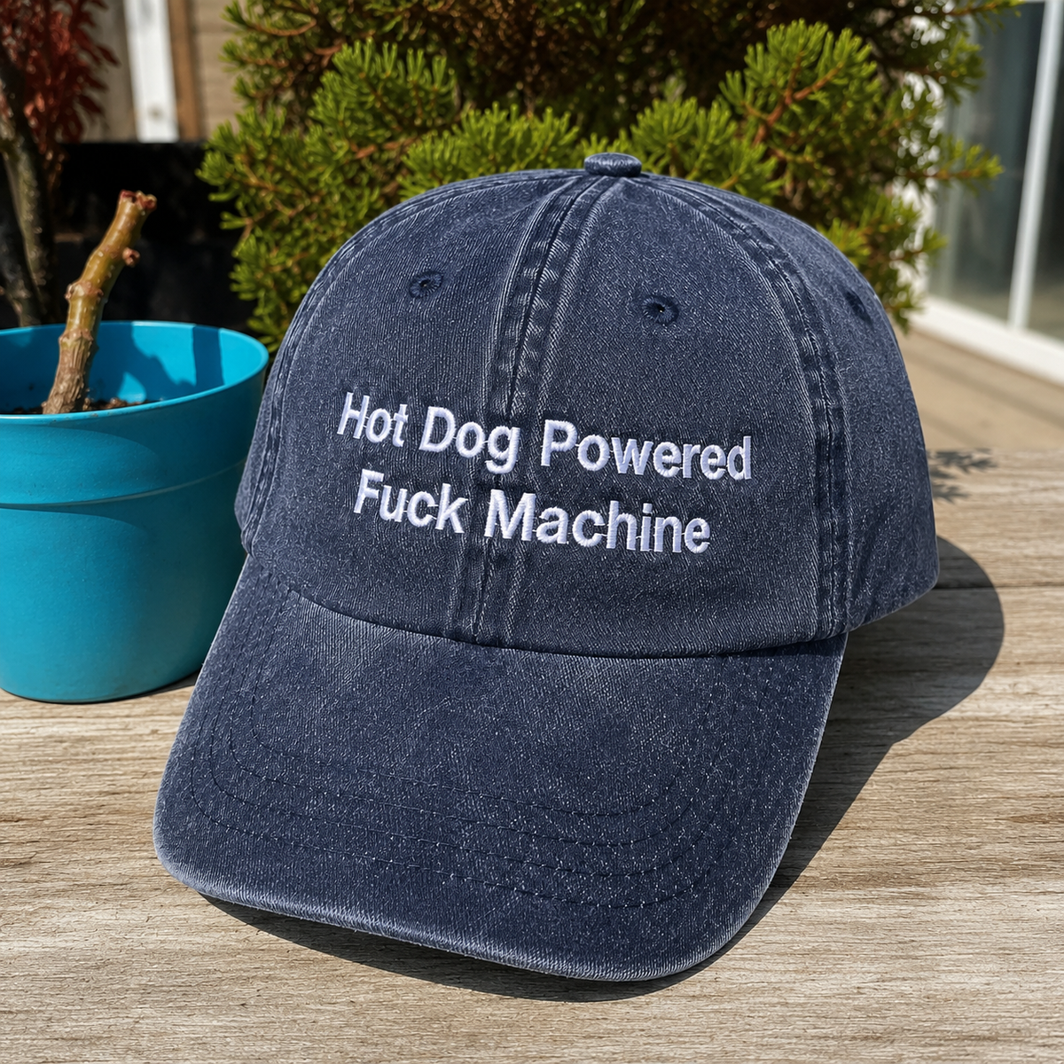 A navy blue washed baseball cap with white embroidered text reading 'Hot Dog Powered Fuck Machine' sits on a wooden deck outdoors next to a blue potted plant.