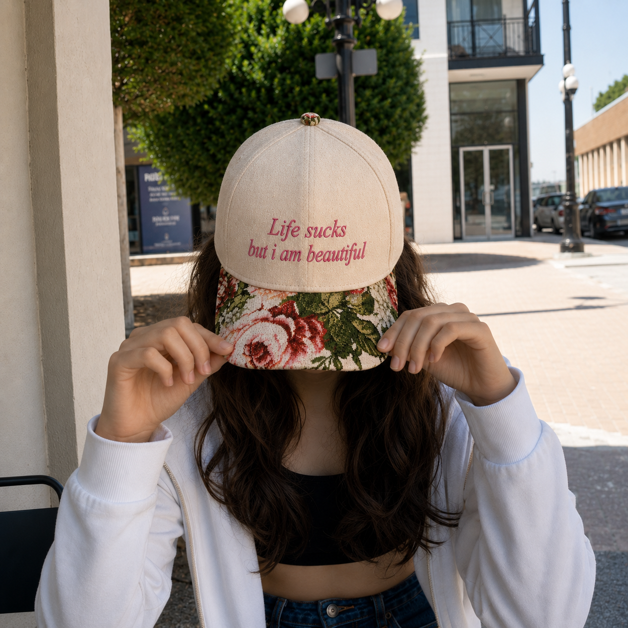 Person wearing a white zip-up hoodie over a black crop top, holding a beige baseball cap embroidered with "Life sucks but i am beautiful" and featuring a floral pattern brim.