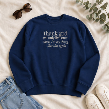 Navy blue embroidered sweatshirt laid flat on a light fabric background, featuring the white quote “thank god we only live once ’cause I’m not doing this shit again,” styled with sunglasses, jeans, white sneakers, and greenery.