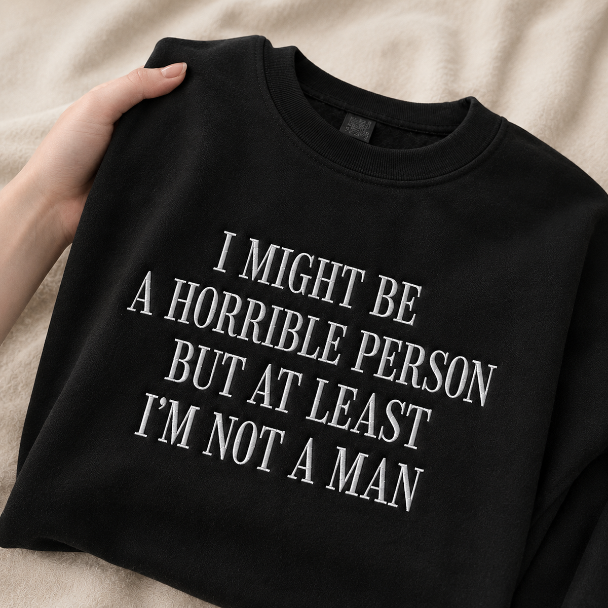 A black crewneck sweatshirt with white text reading 'I MIGHT BE A HORRIBLE PERSON BUT AT LEAST I'M NOT A MAN', held by a hand over a beige textured blanket background.