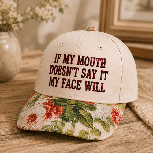 A beige baseball cap with dark red embroidered text reading 'IF MY MOUTH DOESN'T SAY IT MY FACE WILL' on the front, paired with a floral-patterned brim in red, green, and cream, displayed on a wooden surface.