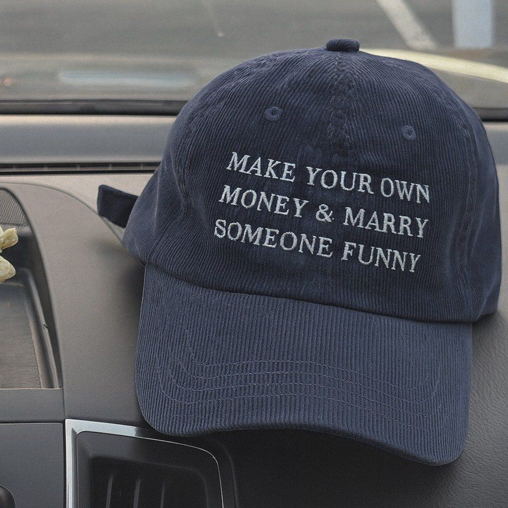A navy blue corduroy baseball cap with white embroidered text reading 'MAKE YOUR OWN MONEY & MARRY SOMEONE FUNNY', resting on a car dashboard.