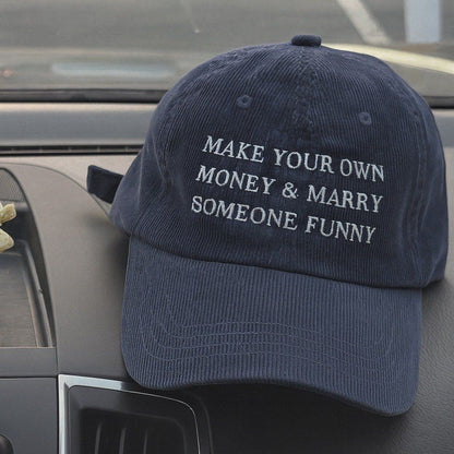 A navy blue corduroy baseball cap with white embroidered text reading 'MAKE YOUR OWN MONEY & MARRY SOMEONE FUNNY', resting on a car dashboard.