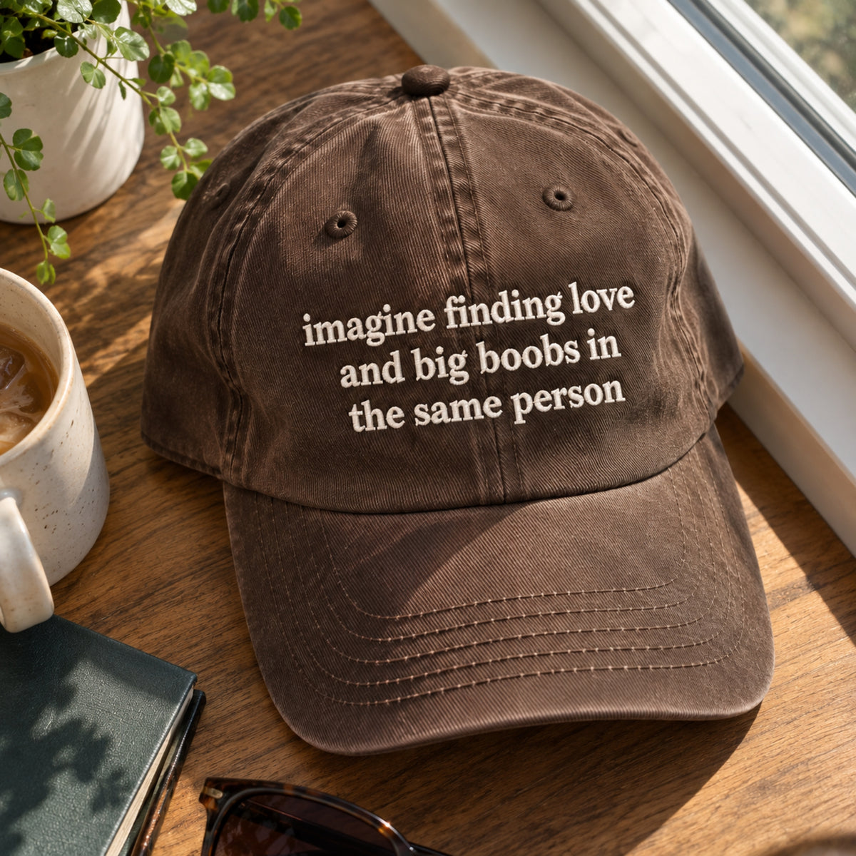 A brown washed baseball cap with white embroidered text reading 'imagine finding love and big boobs in the same person' sits on a wooden table near a window, with a mug, plant, sunglasses, and notebook nearby.