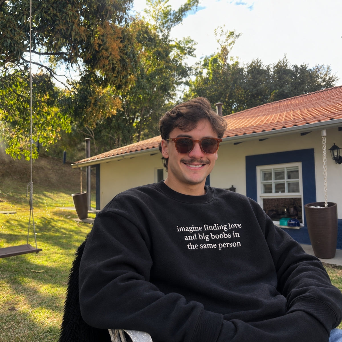 A man wearing a black crewneck sweatshirt with the white printed text 'imagine finding love and big boobs in the same person' sits outdoors in a backyard setting with a house and trees in the background.