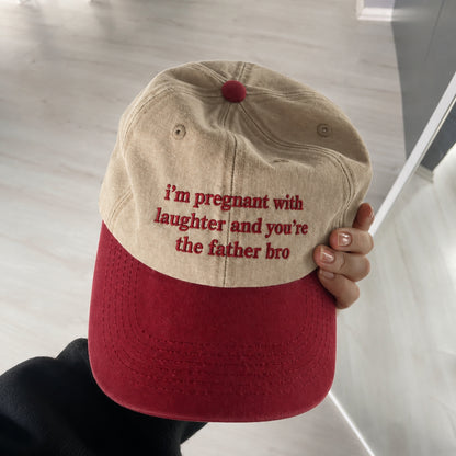 A person holds a dad-style baseball cap with a beige crown and a red brim. The front of the cap has red embroidered text that reads, 'i'm pregnant with laughter and you're the father bro'.