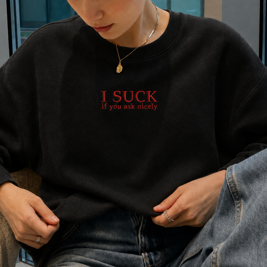 A woman wearing a black crewneck sweatshirt with red text that reads 'I SUCK If you ask nicely' and blue jeans, sitting in a chair in front of large windows.