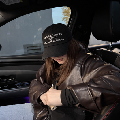A woman sitting inside a car wearing a black baseball cap with white embroidered text that reads 'I SUPPORT A MAN'S RIGHT TO CALL ABORTION EL HOCICO', a brown leather jacket, and a black top.