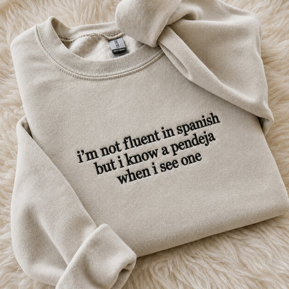 A folded beige crewneck sweatshirt with black printed text reading 'i'm not fluent in spanish but i know a pendeja when i see one', styled on a soft cream faux-fur background.