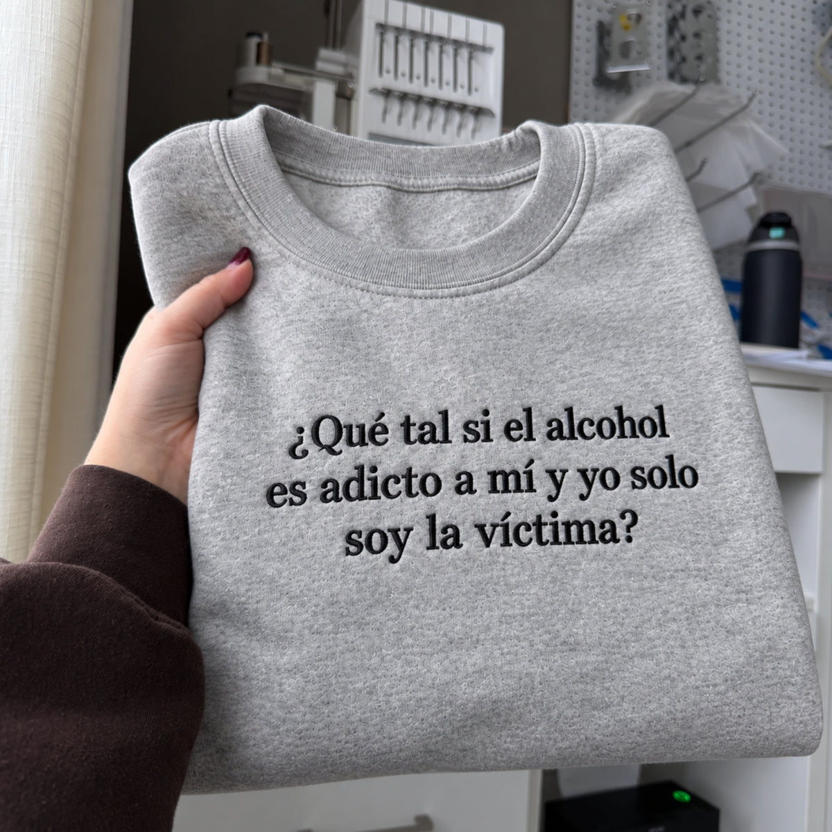 Gray sweatshirt with black embroidered Spanish quote “¿Qué tal si el alcohol es adicto a mí y yo solo soy la víctima?” held up in a casual indoor workspace setting.