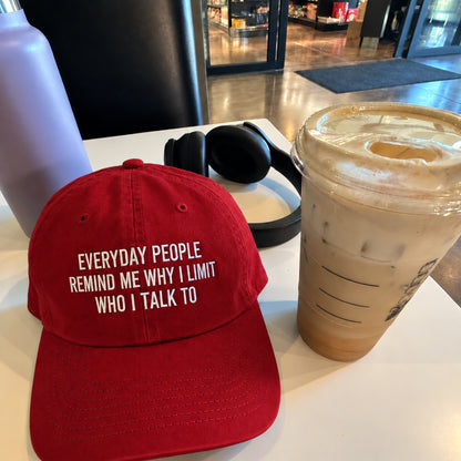 A red baseball cap with white embroidered text reading 'EVERYDAY PEOPLE REMIND ME WHY I LIMIT WHO I TALK TO' sits on a white table next to an iced coffee and black headphones in a cafe setting.