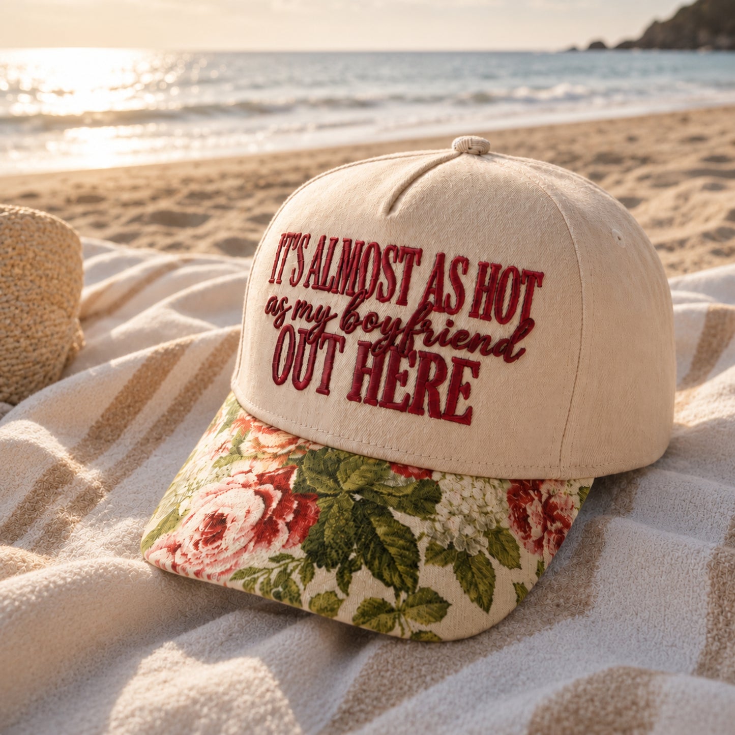 A baseball cap placed on a beach blanket with the ocean and shoreline in the background. The cap is off-white with dark red embroidered text reading “IT’S ALMOST AS HOT as my boyfriend OUT HERE” and has a floral patterned visor in cream, pink, and green.