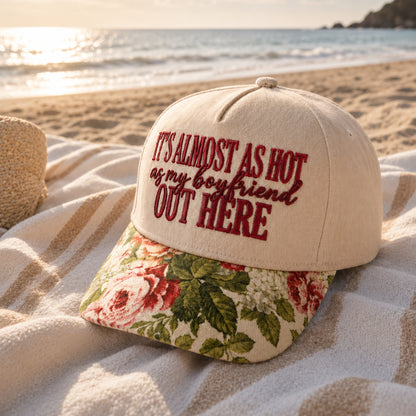 A baseball cap placed on a beach blanket with the ocean and shoreline in the background. The cap is off-white with dark red embroidered text reading “IT’S ALMOST AS HOT as my boyfriend OUT HERE” and has a floral patterned visor in cream, pink, and green.