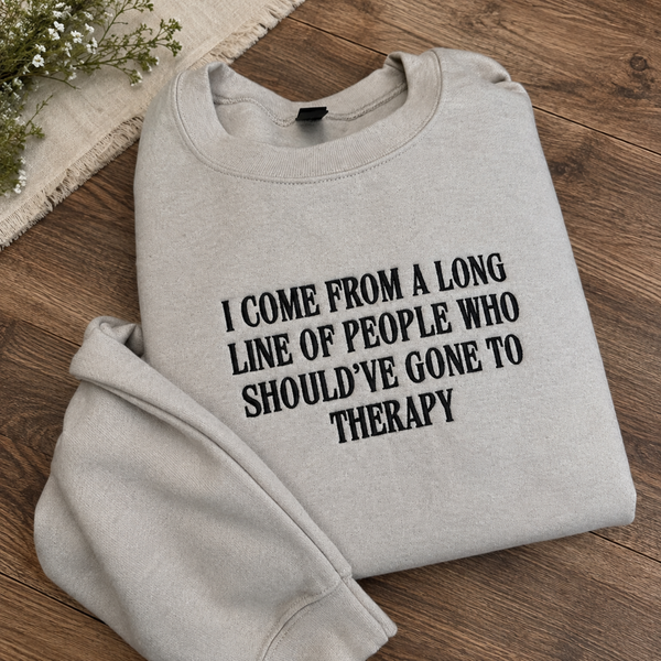 A light gray sweatshirt with black text reading 'I COME FROM A LONG LINE OF PEOPLE WHO SHOULD'VE GONE TO THERAPY' laid flat on a wooden surface with small flowers in the corner.