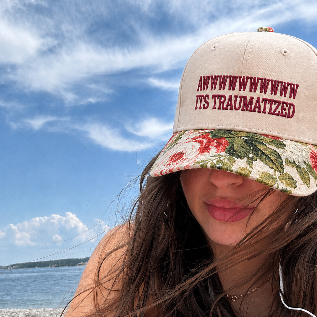 A woman wearing a beige baseball cap with a floral visor and red embroidered text reading 'Awwwwwwwww its traumatized' poses outdoors near the water under a blue sky with clouds.