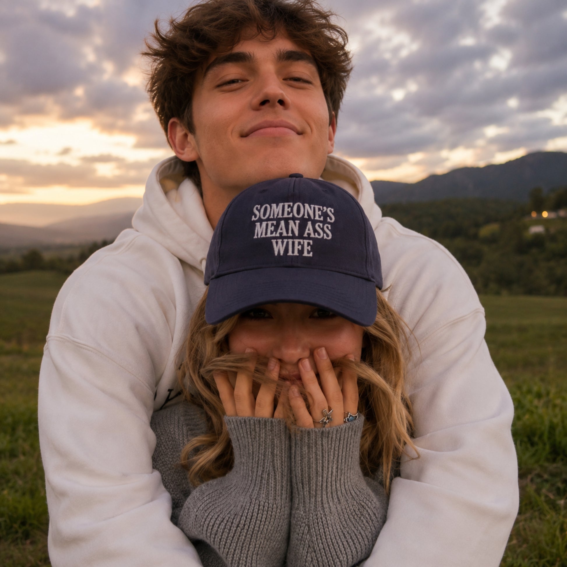 A man in a white hoodie holds a woman in a gray sweater close to his chest outdoors at sunset. The woman is wearing a navy blue baseball cap with the white embroidered text 'SOMEONE'S MEAN ASS WIFE'.