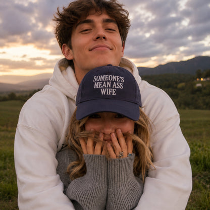A man in a white hoodie holds a woman in a gray sweater close to his chest outdoors at sunset. The woman is wearing a navy blue baseball cap with the white embroidered text 'SOMEONE'S MEAN ASS WIFE'.