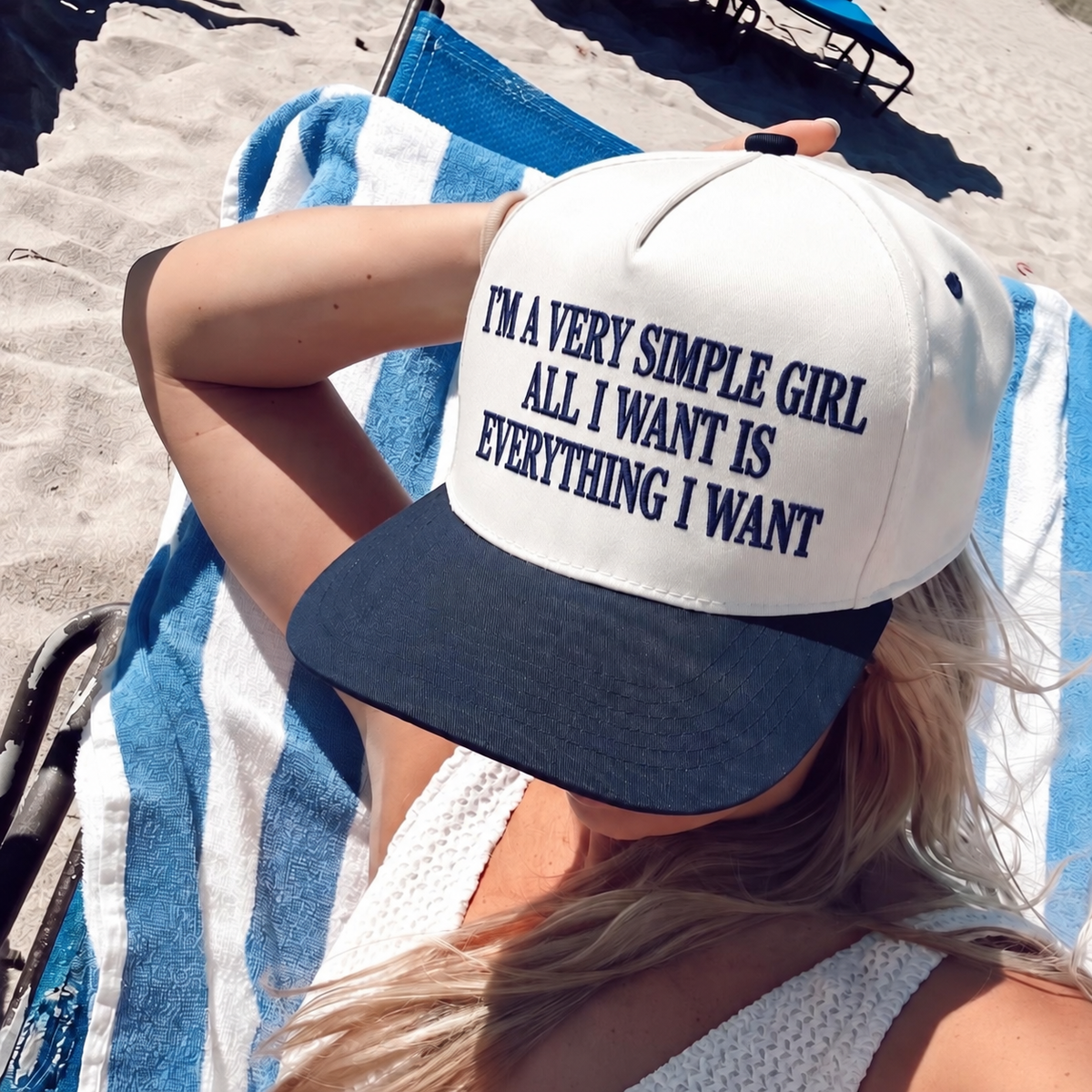 A woman lying on a blue and white striped beach towel on the sand, wearing a white and navy baseball cap with the text 'I'M A VERY SIMPLE GIRL ALL I WANT IS EVERYTHING I WANT'.