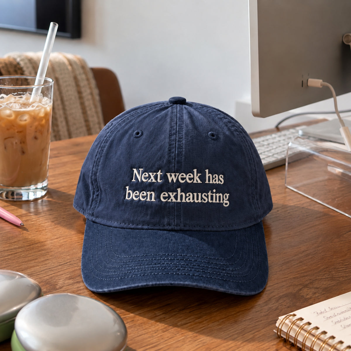 A navy blue baseball cap with the phrase 'Next week has been exhausting' embroidered in white on the front, resting on a wooden desk next to an iced coffee and computer accessories.