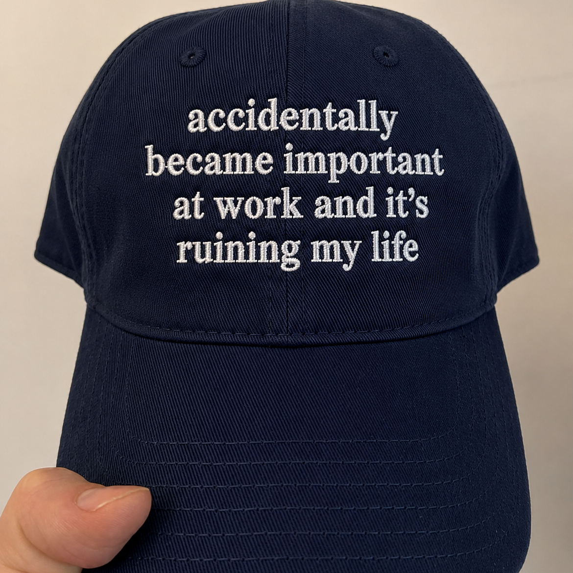 A navy blue baseball cap held by a hand, featuring white embroidered text on the front that reads 'accidentally became important at work and it's ruining my life'.