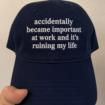 A navy blue baseball cap held by a hand, featuring white embroidered text on the front that reads 'accidentally became important at work and it's ruining my life'.
