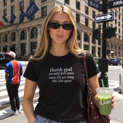 Woman wearing a black embroidered T-shirt with the white quote “thank god we only live once ’cause I’m not doing this shit again,” styled with sunglasses and jeans while holding a green drink on a sunny city street.