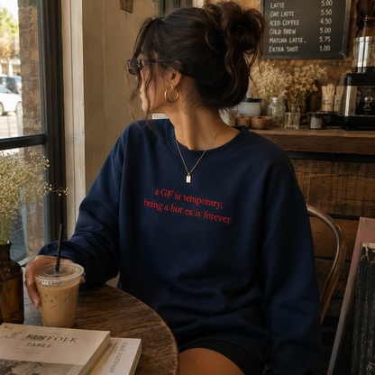 Woman sitting in a coffee shop holding an iced drink, wearing a navy blue crewneck sweatshirt with red embroidered text reading "a GF is temporary, being a hot ex is forever".