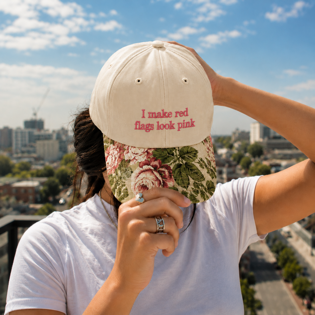 Person holding a beige embroidered baseball hat with pink text reading 'I make red flags look pink' and a vintage floral brim against a sunny sky background.
