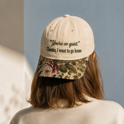 Back view of a woman wearing a beige baseball cap with black embroidered text 'You're so quiet. Thanks, I want to go home.' and a vintage floral tapestry visor