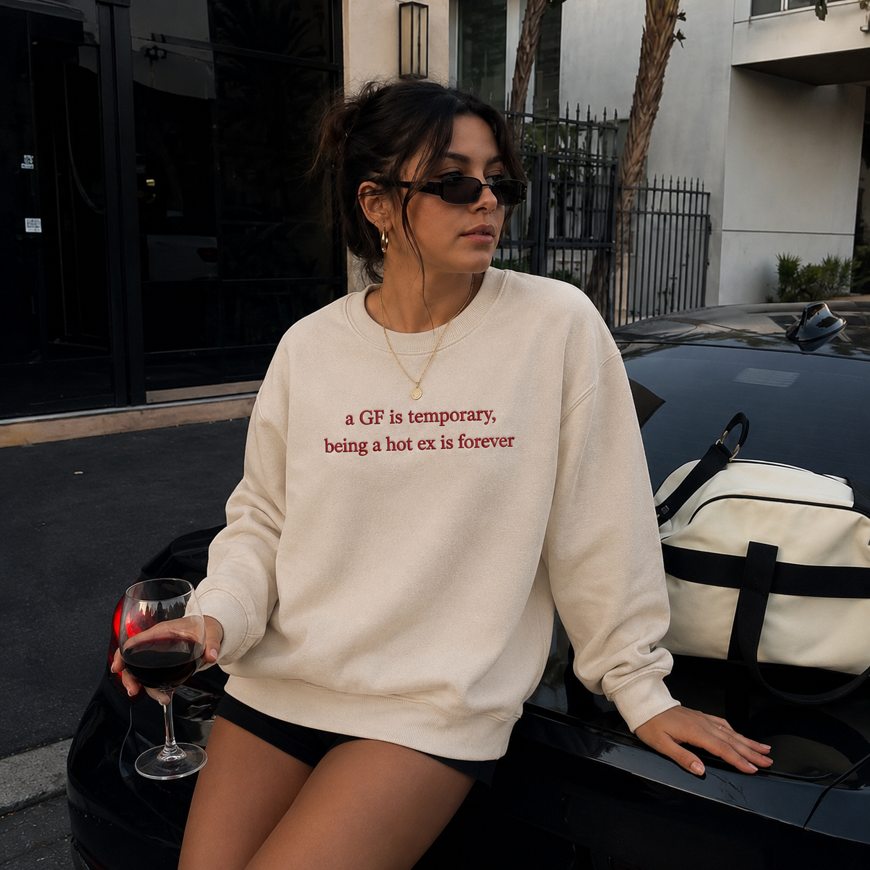Woman wearing sunglasses and a cream-colored crewneck sweatshirt leaning against a car while holding a glass of red wine, embroidered with red text reading "a GF is temporary, being a hot ex is forever".
