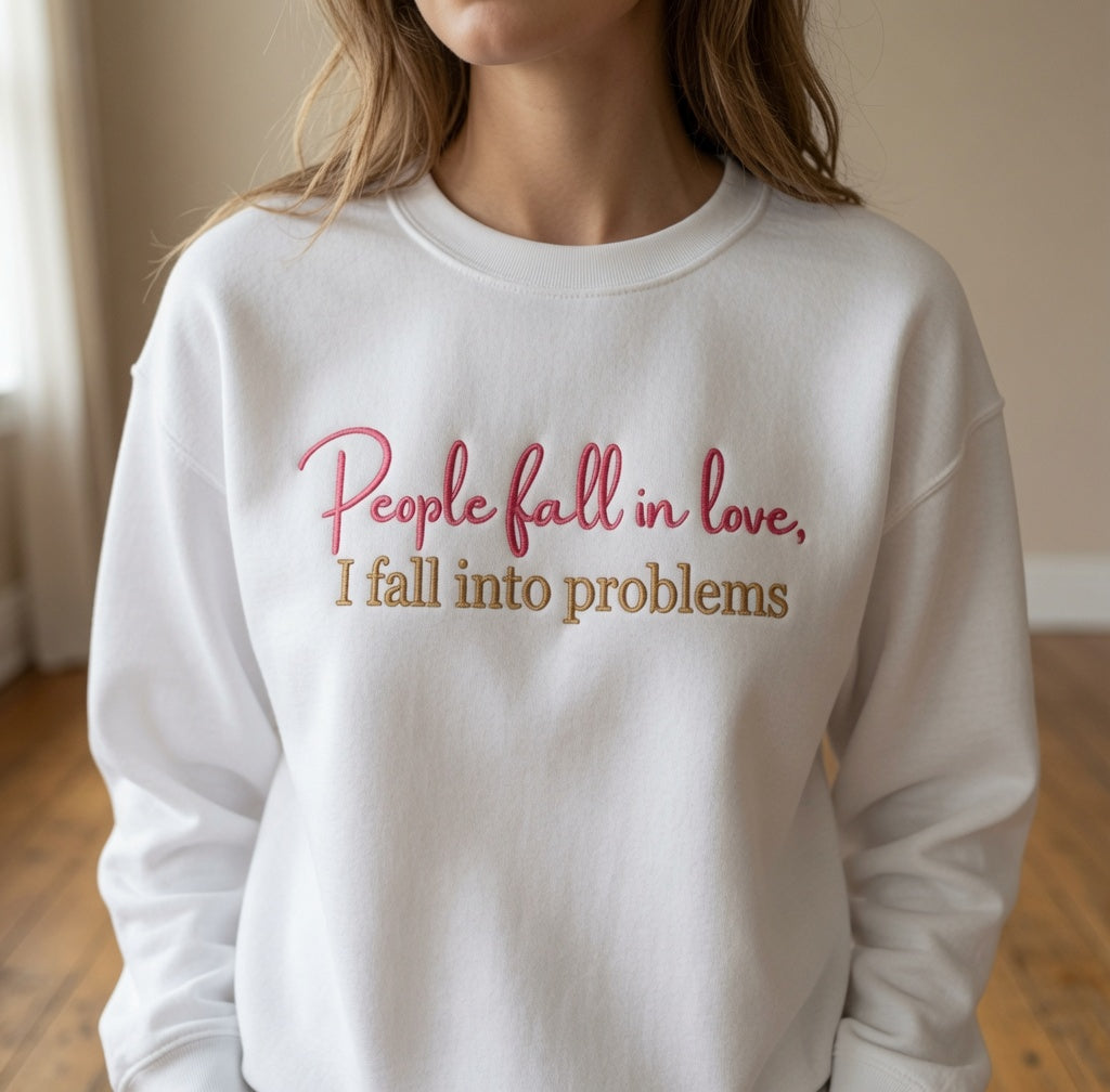 Woman wearing a white crewneck sweatshirt embroidered with pink cursive text reading "People fall in love," above tan block lettering reading "I fall into problems."