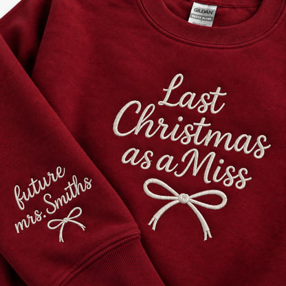 A close-up of a red crewneck sweatshirt with white embroidered text and bows. The right chest reads 'Last Christmas as a Miss' and the left sleeve reads 'future Mrs.