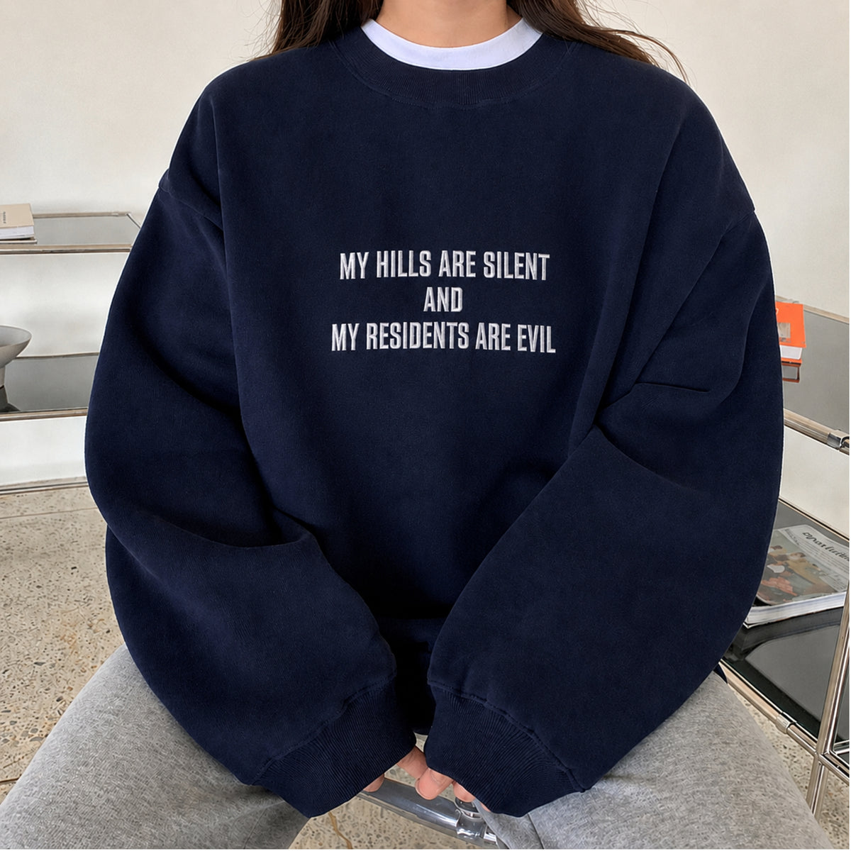 A person wearing an oversized navy blue sweatshirt with white text that reads 'MY HILLS ARE SILENT AND MY RESIDENTS ARE EVIL', paired with light gray sweatpants in an indoor setting.