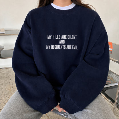A person wearing an oversized navy blue sweatshirt with white text that reads 'MY HILLS ARE SILENT AND MY RESIDENTS ARE EVIL', paired with light gray sweatpants in an indoor setting.