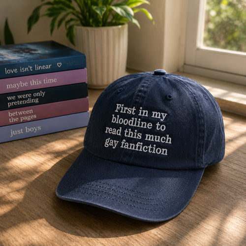 A navy blue baseball cap with white embroidered text reading 'First in my bloodline to read this much gay fanfiction' sits on a wooden table next to a stack of books and a potted plant near a window.