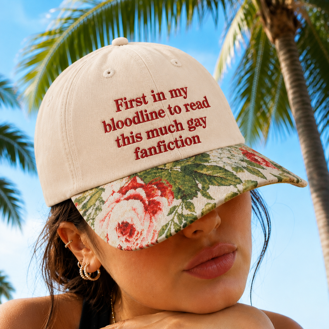 A woman outdoors wearing a beige baseball cap with red embroidered text reading 'First in my bloodline to read this much gay fanfiction' and a floral visor in cream, red, and green. Palm trees and blue sky are visible in the background.