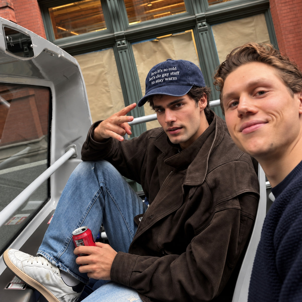 Two men sitting inside the open trunk of a vehicle, with one wearing a dark brown jacket, blue jeans, and a navy blue baseball cap with white text, while the other man is in the foreground.