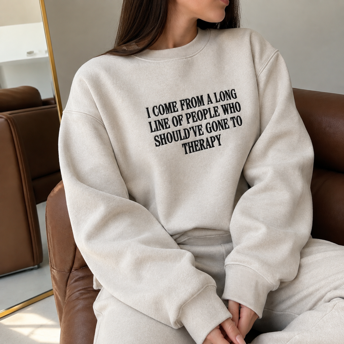 A woman wearing a light beige crewneck sweatshirt with black text that reads 'I COME FROM A LONG LINE OF PEOPLE WHO SHOULD'VE GONE TO THERAPY', sitting on a brown leather couch.