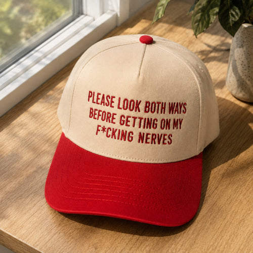A red and beige baseball cap with the text 'PLEASE LOOK BOTH WAYS BEFORE GETTING ON MY F*CKING NERVES' embroidered in red on the front, resting on a wooden windowsill.