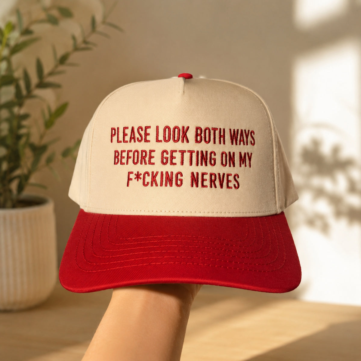 A beige and red trucker-style cap held up by a hand, featuring the humorous text 'PLEASE LOOK BOTH WAYS BEFORE GETTING ON MY F*CKING NERVES' in red embroidery on the front panel. The background is a warm, sunlit indoor setting with a blurred potted plant.