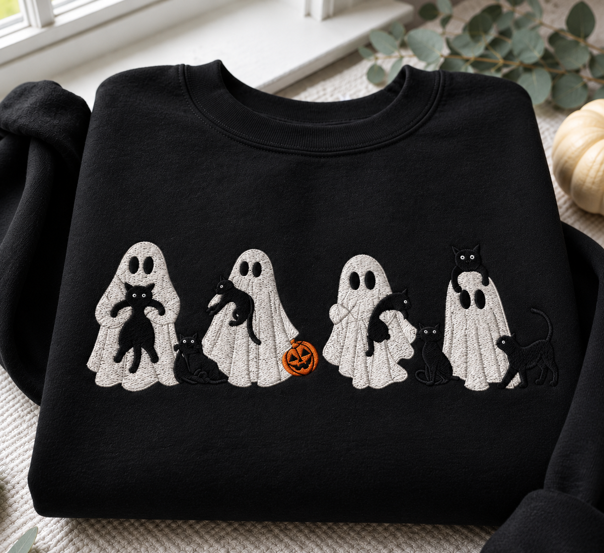 A black crewneck sweatshirt featuring a horizontal embroidered design of five white ghosts holding or standing with black cats and a small orange pumpkin, displayed on a neutral textured surface with seasonal props.