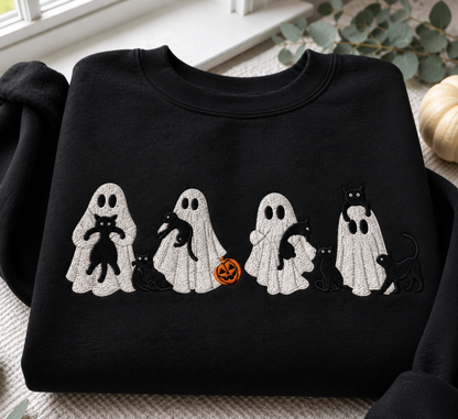 A black crewneck sweatshirt featuring a horizontal embroidered design of five white ghosts holding or standing with black cats and a small orange pumpkin, displayed on a neutral textured surface with seasonal props.
