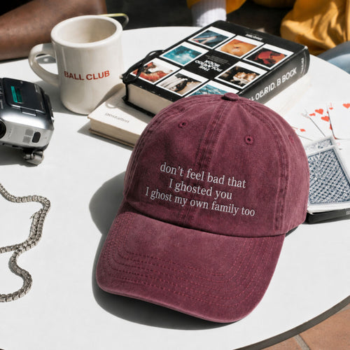 A dusty burgundy baseball cap with white embroidered text reading 'don't feel bad that I ghosted you I ghost my own family too' sits on a white table surrounded by lifestyle props including a mug, camera, book, and cards.