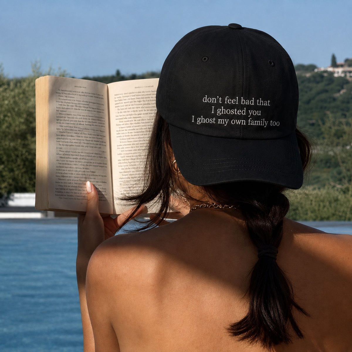 A woman seen from behind outdoors by a pool, wearing a black baseball cap with white embroidered text that reads, 'don't feel bad that I ghosted you I ghosted my own family too' while holding an open book.